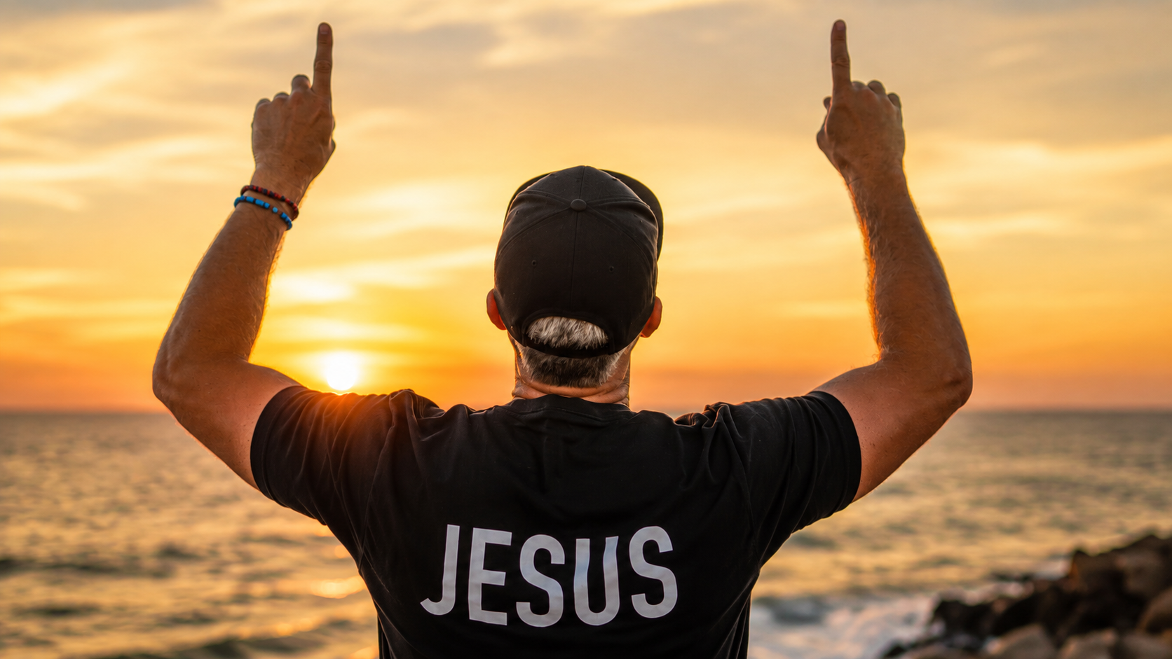 Founder standing on coastal rocks at sunset with arms outstretched wearing a black THE ANSWER IS... tee with JESUS in white Impact on the back