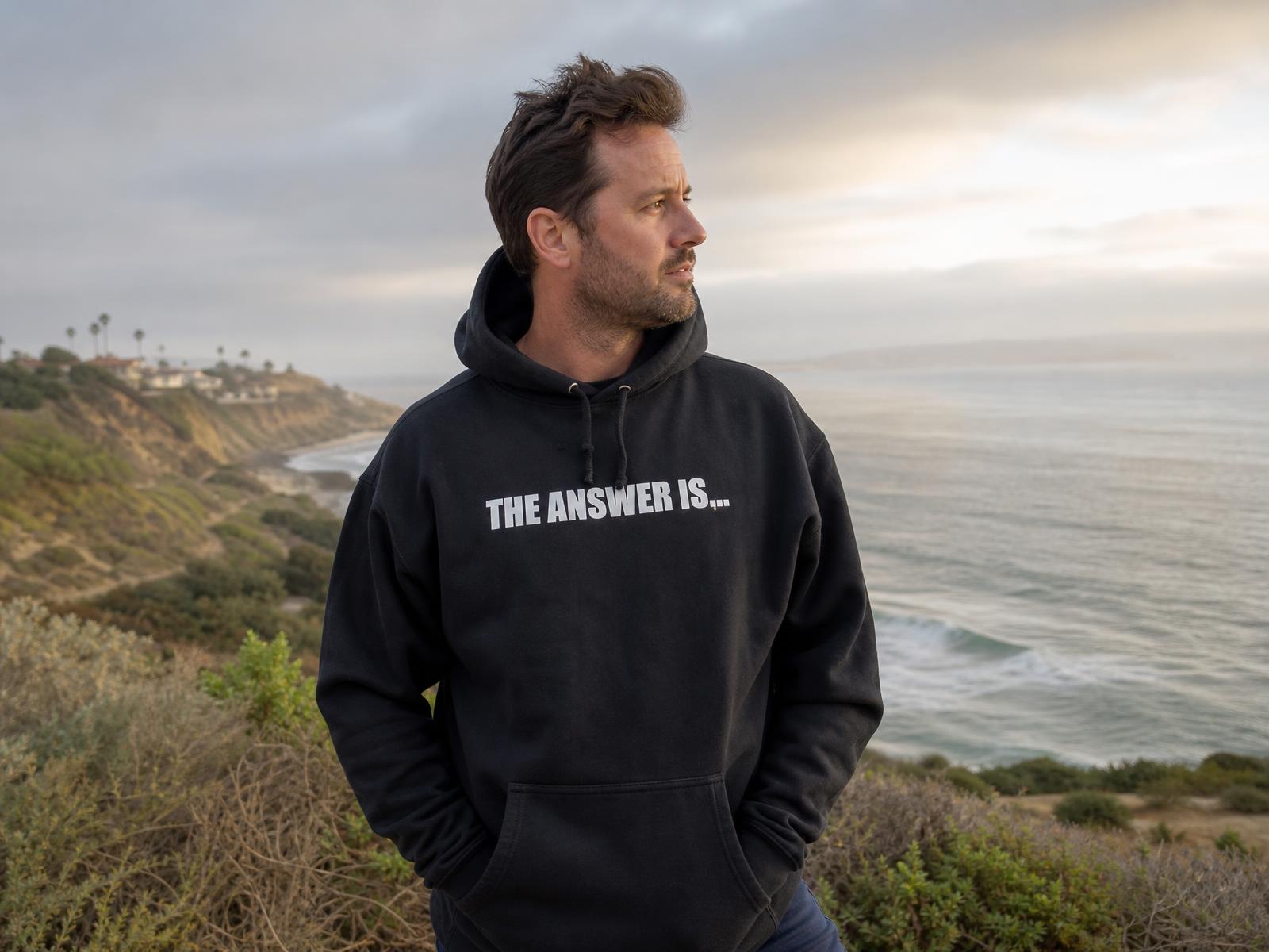 Man in a black THE ANSWER IS... pullover hoodie standing at a Southern California coastal overlook in cool early-morning marine-layer light.